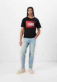 Zwarte katoen T-shirt met rood en wit "JACK & JONES" graphic, gecombineerd met lichtblauwe versleten jeans en beige sneakers.