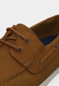 Καφέ boat shoe από καστόρι με δερμάτινα κορδόνια περασμένα μέσα από μεταλλικά ρουξούνια και λευκή σόλα ορατή στο κάτω μέρος.