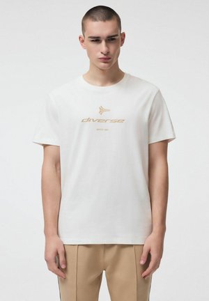 Młody mężczyzna w białej koszulce z beżowym logo "diverse" i napisem "SINCE 1993", w beżowych spodniach, stojący na jednolitym tle.