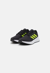 Baskets de sport noires avec une tige en maille texturée, présentant trois rayures vert fluo. La semelle est blanche avec un accent noir sur l'orteil.