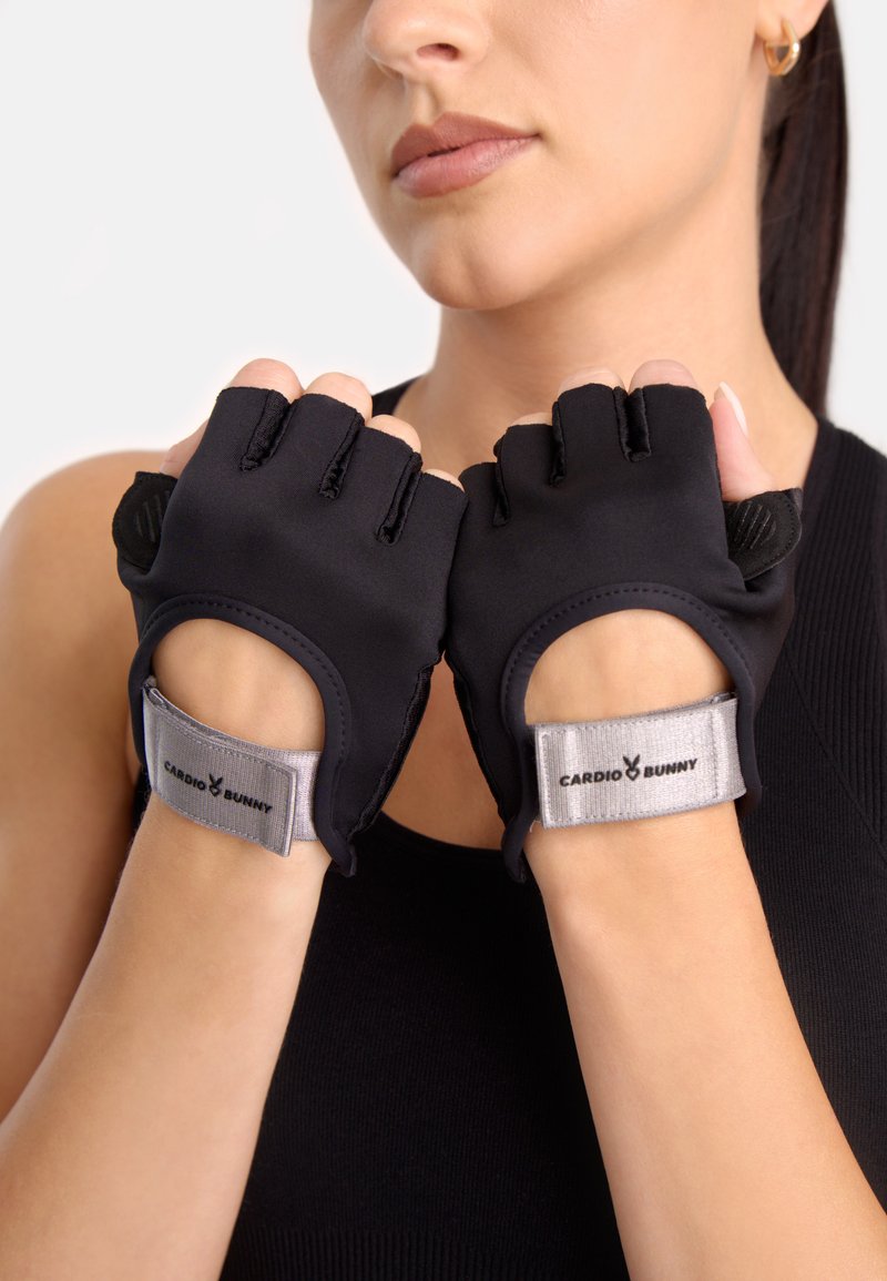 Mujer con guantes negros para hacer ejercicio sin dedos y correas grises en las muñecas con la etiqueta "Cardio Bunny", con las manos cerca de su rostro.