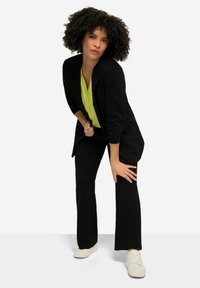 Blazer noir ajusté sur un chemisier vert vif, associé à un pantalon noir évasé et des baskets blanches. Cheveux bouclés et fond neutre.