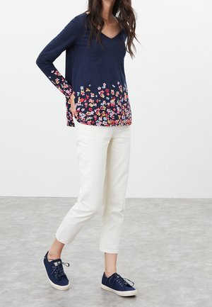 Navy V-neck long-sleeve top with floral pattern at the hem, paired with white cropped pants and navy lace-up sneakers.