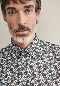 Bluse mit floralem Muster auf braunem Grund, verziert mit weißen und blauen Blumen. Knopfleiste und Kragen, aus weichem Stoff gefertigt.
