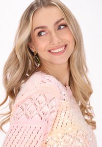 Pleteni pulover u pastelno roza boji s prozornim dizajnom i šljokicama. Model nosi zlatne kružne naušnice, pokazuje valovitu kosu i blistav osmijeh.