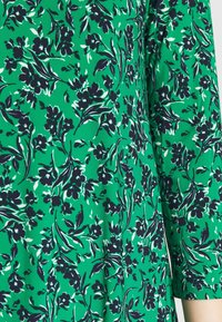 Green floral patterned fabric featuring dark blue flowers and leaves, with a smooth texture and short sleeves.