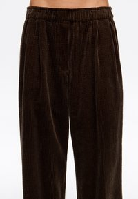 Pantalones de pana marrón con cintura elástica, llevados por una persona, mostrados desde la cintura hasta la mitad del muslo sobre un fondo blanco liso.