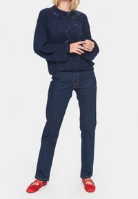 Pull en maille bleu marine orné de sequins, manches ballon, associé à un jean bleu foncé et des chaussures plates rouges à nouer.