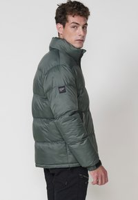 Chaqueta tipo puffer en verde oscuro, con un alto cuello, diseño acolchado, parche de acento negro en la manga y puños acanalados para mayor calidez.