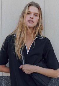 Schwarzes kurzärmliges Poloshirt mit einem weißen Kragen, mit einem V-Ausschnitt-Design. Das Modell hält eine schwarze Tasche vor einem neutralen Hintergrund.