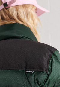 Giacca verde e nera con una texture liscia, caratterizzata da un pannello spalla nero a contrasto e un'etichetta "SUPERDRY" sul bordo.