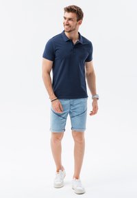 Mörkblå polo med krage, kombinerad med ljusblå denimshorts. Modellen har på sig vita sneakers med orange detaljer.