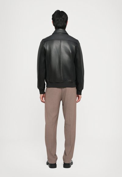 Zwarte leren bomberjack met ribbed manchetten en zoom, met een recht gesneden ontwerp. Draagt bij-beige tailored broek en zwarte schoenen.