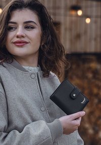 Black leather wallet with a snap closure, featuring a logo. Smooth texture, rectangular shape. Model wears a gray bomber jacket.