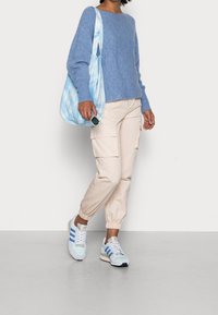 Pull en maille bleu clair, pantalons cargo beiges avec poignets élastiques et baskets pastel. Accessoirisé d'un sac fourre-tout tie-dye et de lunettes de soleil.