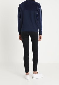 Mörkblå, mjuk långärmad sweatshirt med en slät, kontrasterande tygpanel på övre ryggen, kombinerad med tajta svarta jeans och vita sneakers.