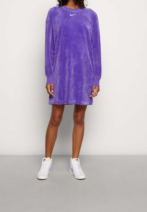 Osoba w fioletowej, oversize'owej sukience z korduru marki Nike i białych, wysokich trampkach Nike, stojąca na tle jednolitego, jasnego tła.