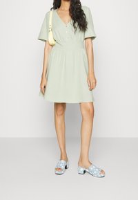 Máta zelené šaty s krátkým rukávem, V-výstřihem a detaily knoflíků, spárované s modrými květinovými dřeváky s nízkým černým podpatkem.