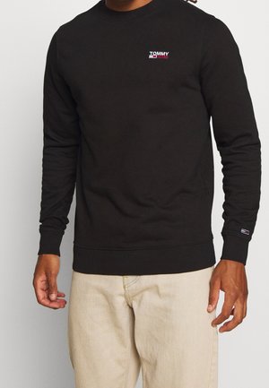 Homme portant un sweat-shirt noir Tommy Jeans et un pantalon beige, debout avec les mains détendues le long du corps sur un fond uni.