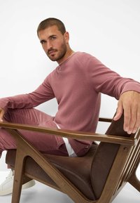 Rosa strukturiertes Sweatshirt mit gerippten Bündchen, kombiniert mit passenden Hosen. Modell sitzt auf einem Holzstuhl mit einem braunen Ledersitzkissen.