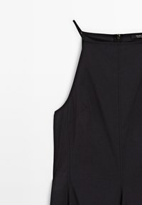 Černý overal s tenkými ramínky, s vypasovaným střihem, bočními švy a zipem na zádech. Vyroben z hladké tkaniny s minimální texturou.