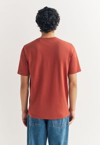 Puha pamutból készült, rozsdabarna rövid ujjú póló, klasszikus kerek nyakkivágással és laza szabással, kék farmernadrággal párosítva.