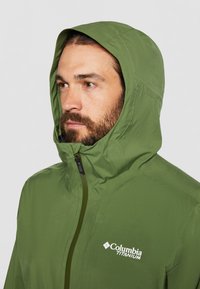 Hombre con barba que lleva una chaqueta con capucha verde Columbia Titanium abrochada hasta la barbilla, mirando ligeramente hacia la izquierda sobre un fondo liso.