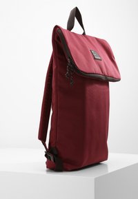 Mochila de tela burdeos con solapa plegable, acentos de cremallera y un asa marrón. Presenta una forma rectangular con una textura suave.