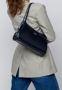 Sac en cuir navy au toucher lisse, de forme arrondie, avec des ferrures en argent. Porté à l'épaule, associé à un blazer à carreaux.
