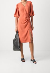 Robe couleur rouille avec un design en portefeuille, manches courtes bouffantes, fronces sur le côté et fente haute sur la cuisse. Associée à un cabas en cuir noir et des mules ouvertes.