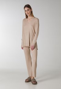 Beige geribbelde trui met V-hals en twee zakken, gecombineerd met bijpassende geribbelde broeken en bruine slippertjes. Zachte textuur en een relaxte pasvorm.