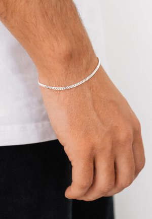 Sølv kædearmbånd med et fladt, tekstureret design, båret på et håndled. Kæden har indbyrdes låste led og en glat finish.