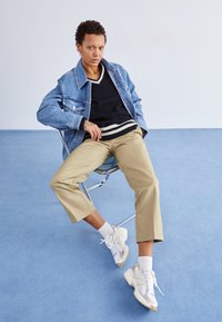 Person med kort lockigt hår bär en denimjacka, svart tröja, beige byxor och vita sneakers, sitter avslappnat på en hopfällbar stol på en blå golv.