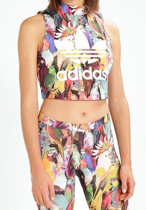 Mouwloze crop top en legging met kleurrijk vogelpatroon en groot wit Adidas-logo op de top, gedragen door een vrouw.