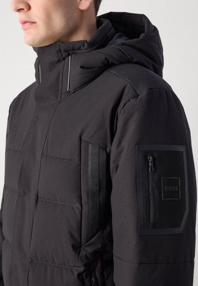 Veste matelassée noire avec capuche, dotée de poches zippées, d'un patch logo en relief et d'accents texturés contrastants sur le devant.
