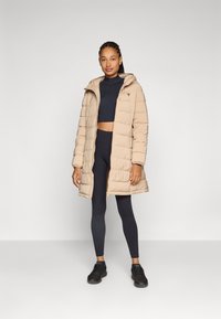 Beige puffer jakke med hette, med quiltet tekstur og glidelås. Båret over en marineblå crop-top og svarte leggings med svarte sko.
