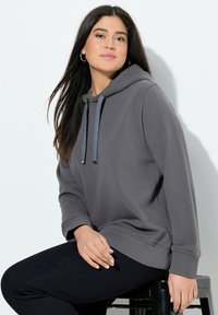 Sudadera gris con cordón, puños y dobladillo acanalados. Tela suave, corte relajado. Usada con jeans oscuros, sentado en un taburete negro.