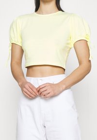 Lysegul cropped top med puffede ærmer, elastikker ved manchetten og rund halsudskæring. Kombineret med hvide bukser med høj talje.