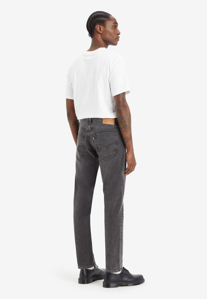 Jean noir avec un délavage gris tonifié, poches arrière avec des détails de couture, porté avec un t-shirt blanc ajusté et des chaussures noires.