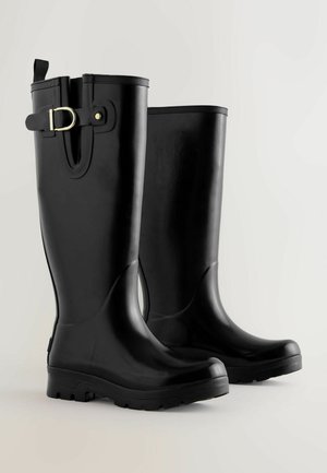 Egy pár magas fekete gumicsizma oldalsó csattal és texturált talppal, világos, egyszínű háttéren.