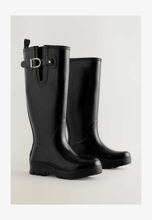 Egy pár magas fekete gumicsizma oldalsó csattal és texturált talppal, világos, egyszínű háttéren.