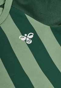 Grön och ljusgrön randig tyg har ett texturerat mönster med en liten vit bi-logotyp i övre vänstra hörnet.