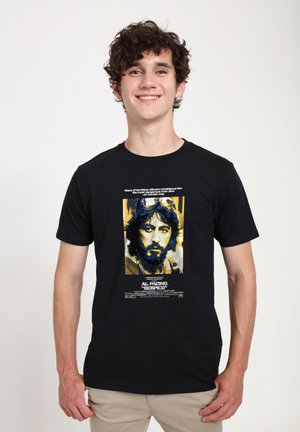 Černé tričko s grafickým potiskem portrétu se žlutými akcenty. Text zní "Al Pacino 'Serpico'" nad a pod obrázkem.