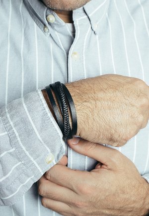 Black leather bracelets in varying textures, including smooth and braided, stacked on a wrist. Gray striped shirt partially visible.