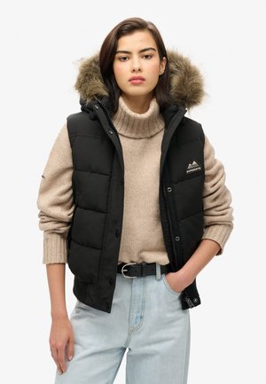 Gilet noir rembourré avec une capuche en fausse fourrure amovible, arborant un patch logo, associé à un pull en laine beige et un jean bleu clair.