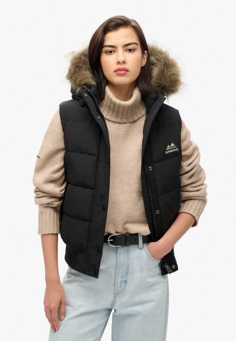 Schwarze Puffer-Weste mit abnehmbarem Kunstpelzkragen, ausgestattet mit einem Logopatch, kombiniert mit einem beigen Strickpullover und hellblauen Jeans.