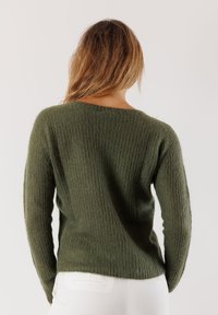 Una donna con capelli lunghi indossa un maglione verde oliva texture e pantaloni bianchi, voltata di spalle contro uno sfondo chiaro e semplice.