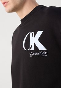 Gros plan d'une personne portant un sweat-shirt noir avec de grands textes blancs "CK" et "Calvin Klein EST 1968" sur la poitrine.