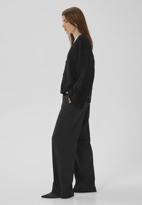 Sieviete stāv melnās plaša griezuma biksēs, nēsā vaļīgu melnu džemperi, rokas kabatās, raugās uz sāniem pret vienveidīgu pelēku fonu.