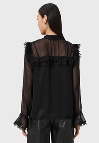 Schwarze durchsichtige Bluse mit Rüschendetails aus Spitze an Schultern und Ärmelbündchen. Lange Ärmel, strukturiertem Stoff und gewelltem Saum.